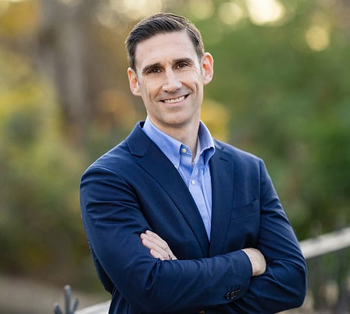 Headshot of Nick Barringer, human performance consultant and executive nutritionist, in a suit coat and button up smiling with his arms crossed